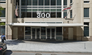 Dr. Patel's Washington DC Office building entrance with the address number 300 displayed prominently above glass doors, featuring contemporary architecture with large windows, stone columns, American and state flags, and a paved plaza area with a pedestrian walking by